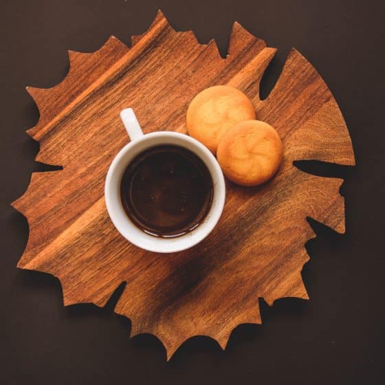 cup of coffee with two cookies on a maple leaf shaped cutting board against a brown background