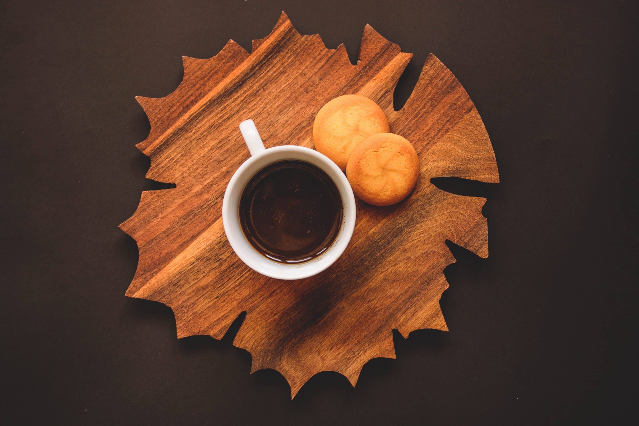 cup of coffee with two cookies on a maple leaf shaped cutting board against a brown background